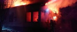 Na zdjęciu w porze nocnej widoczny strażak gaszący pożar budynku mieszkalnego. Warunki zimowe, śnieg, strażak podaje prąd wody w natarciu na palący się dom.