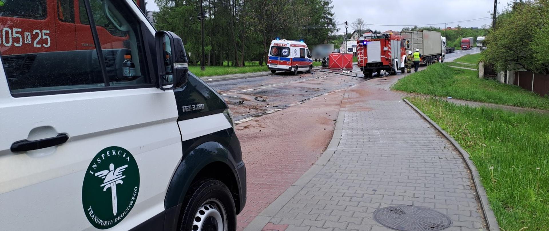 Na pierwszym planie (od lewej): prawy bok oznakowanego furgonu małopolskiej Inspekcji Transportu Drogowego. W tle: karetka pogotowia ratunkowego, wóz straży pożarnej, rozbity samochód osobowy na środku drogi i zestaw ciężarowy biorący udział w wypadku.