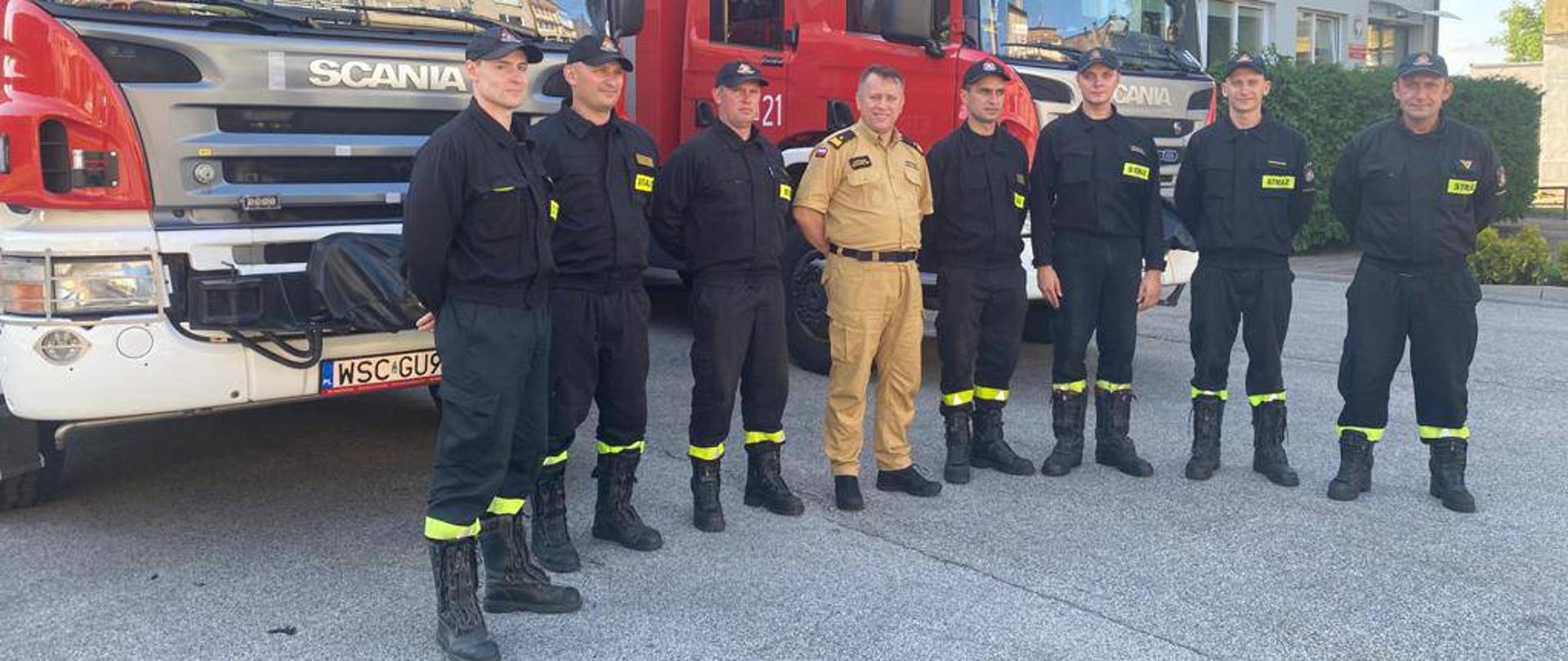 Strażacy sochaczewskiej Jednostki Ratowniczo-Gaśniczej stoją w szeregu, ubrani w czarne ubrania koszarowe. Wśród nich - czwarty o lewej strony, w piaskowym ubraniu, stoi mazowiecki komendant wojewódzki PSP. W tle dwa samochody gaśnicze oraz budynek jednostki.