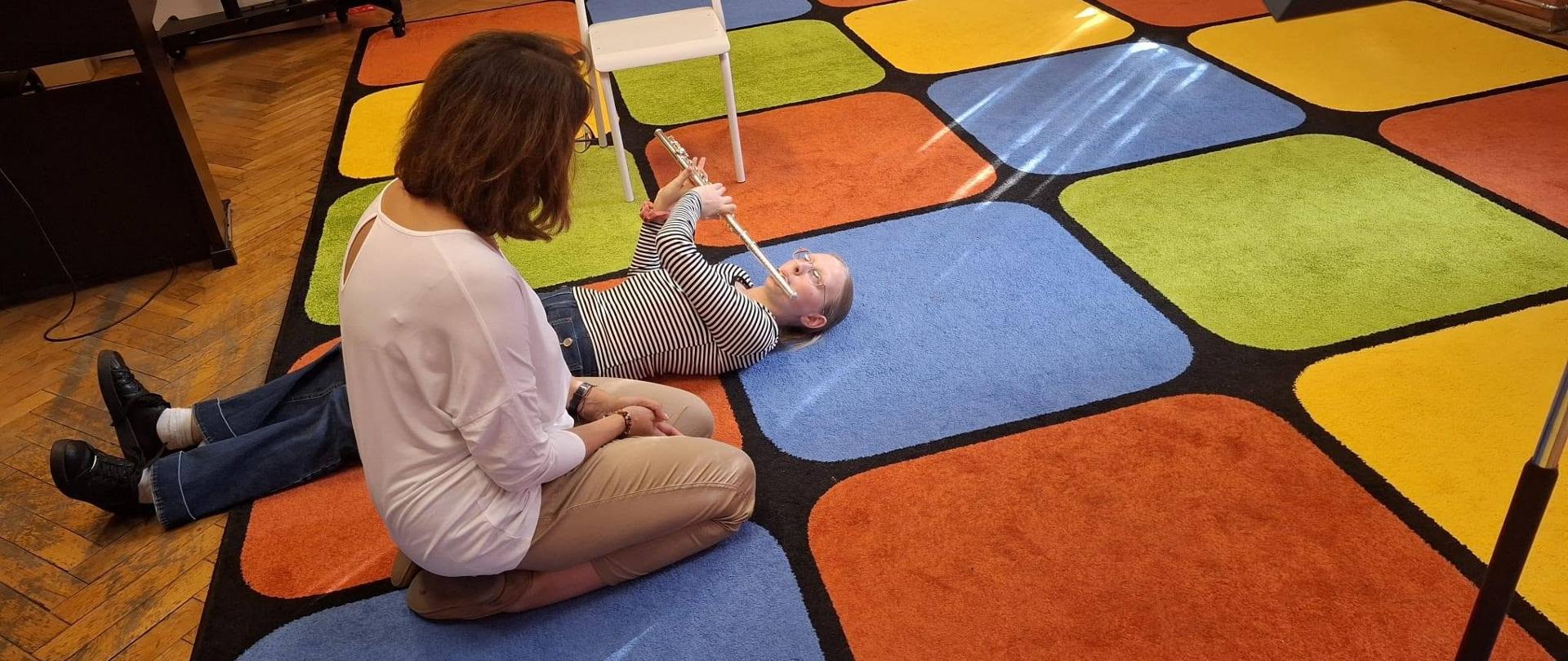 Na kolorowej wykładzinie leżąca uczennica z fletem. Obok niej klęczy nauczyciel