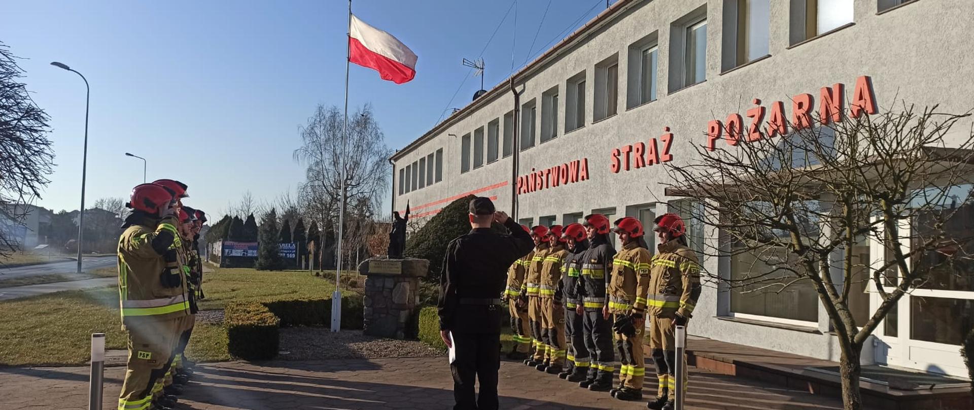 Zdjęcie przedstawia zmianę służby Jednostki Ratowniczo-Gaśniczej w asyście Zastępcę Dowódcy Jednostki Ratowniczo-Gaśniczej, którzy znajdują się przy maszcie flagowym. Przed nimi znajdują się ustawione do siebie równolegle zmiana zdająca i przyjmująca służbę, ubrani w odzież specjalną z hełmami. W tle widać napis "Państwowa Straż Pożarna" na budynku komendy, drzewa.