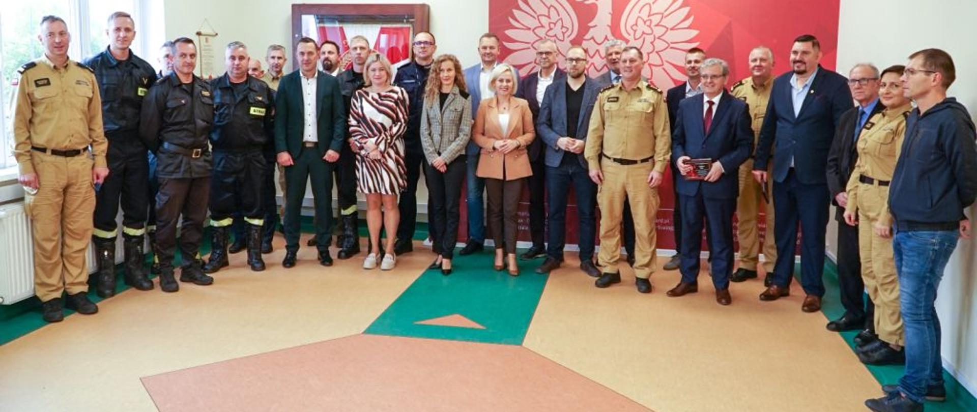 Zdjęcie grupowe wykonane na świetlicy Komendy PSP w Białogardzie. Mężczyźni , kobiety, strażacy, cywile, stoją przed ścianką reklamową KP PSP Białogard. Ścianka czerwona, z logiem PSP.