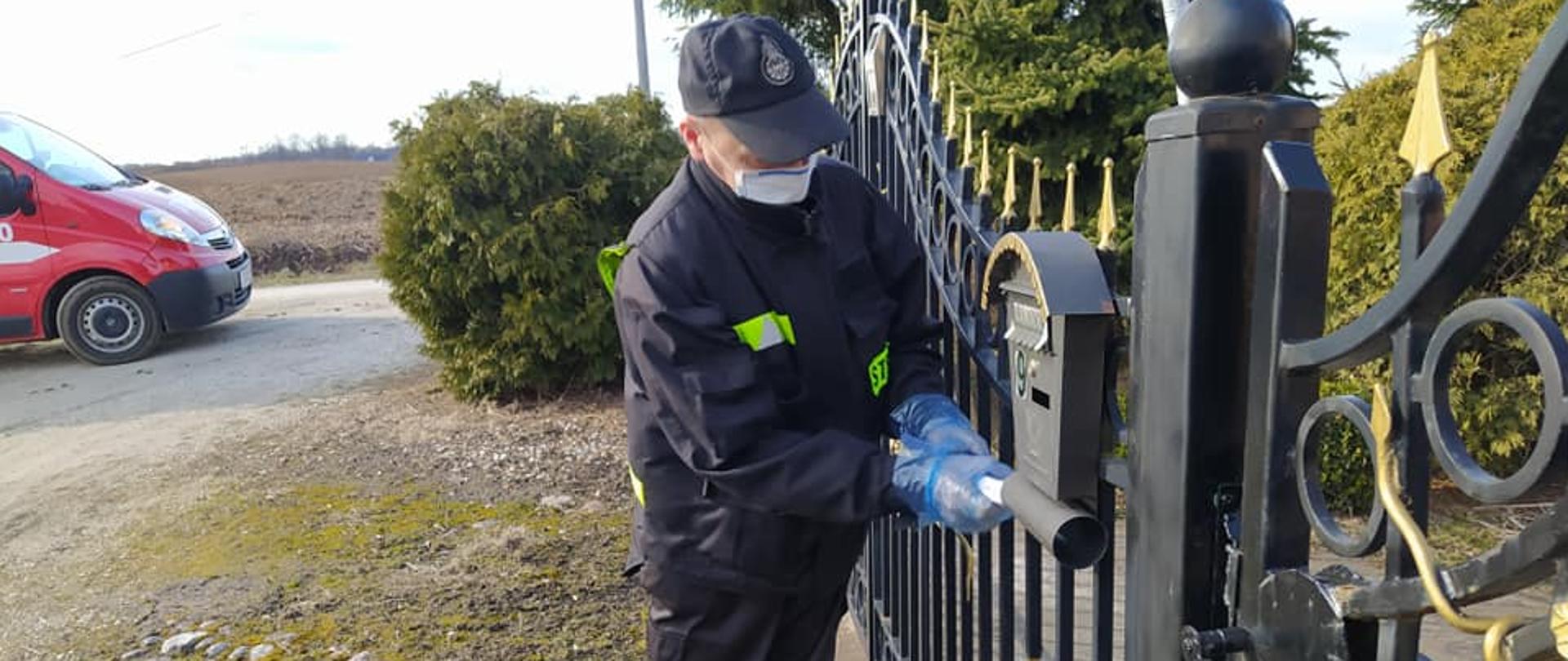 NA ZDJĘCIU WIDAĆ MĘŻCZYZNĘ STRAŻAKA, KTÓRY WKŁADA ZWINIĘTĄ ULOTKĘ INFORMACYJNĄ DO SKRZYNKI POCZTOWEJ. W TLE WIDAĆ FRAGMENT SAMOCHODU OSP