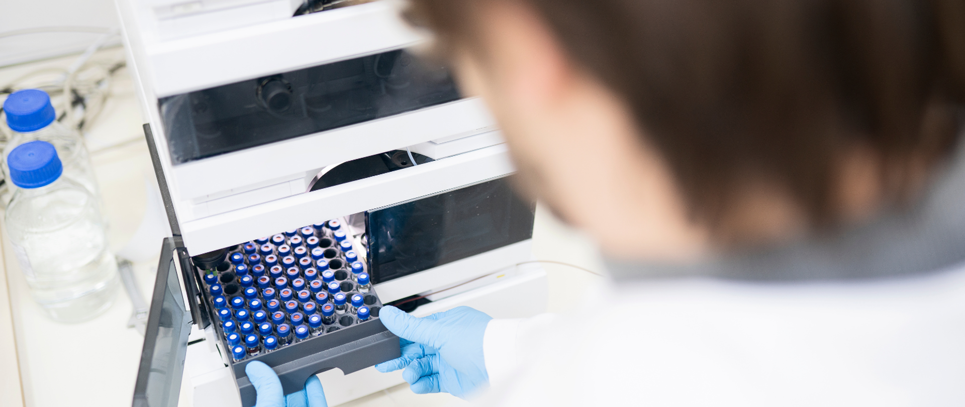 Pracownik laboratorium w rękawiczkach ochronnych wkłada do urządzenia analitycznego tacę z próbkami w fiolkach.