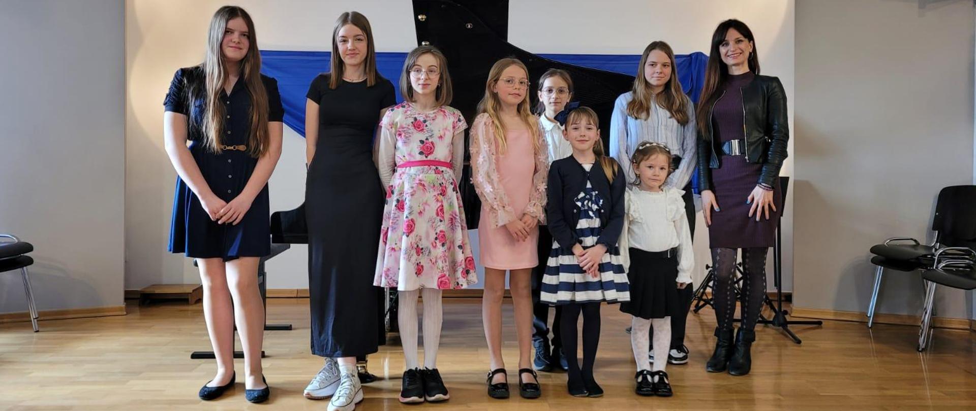 Na zdjęciu grupa uczniów. Same dziewczynki. Jedne starsze, inne dużo młodsze. Po prawej stronie zdjęcia kobieta brunetka. Wszyscy stoją na drewnianym parkiecie. w tyle za nimi czarny fortepian, a na ścianie niebieski materiał. Po prawej stronie brązowe krzesła na srebrnych nogach.