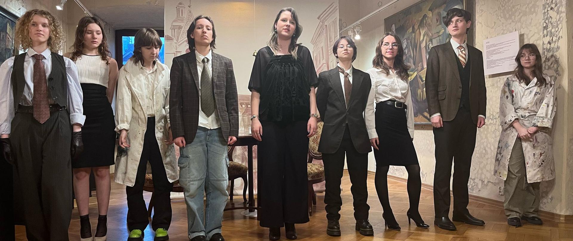 Młode osoby w różnych przebraniach (eleganckich ubraniach, białych fartuchach poplamionych farbą), stoją w jednym rzędzie, przodem do obserwatora, w przestrzeni muzealnej. Za nimi stół, na ścianach obrazy