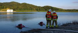 Widok z tyłu. Dwóch strażaków w ubraniach specjalnych stoi nad wodą, w której widoczne są dwie motopompy pływające.