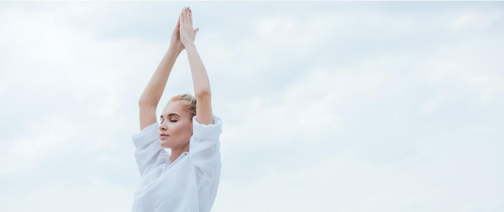 Na zdjęciu jest kobieta z uniesionymi nad głowom rękoma, które są złożone. Tło jest w jasnych kolorach.