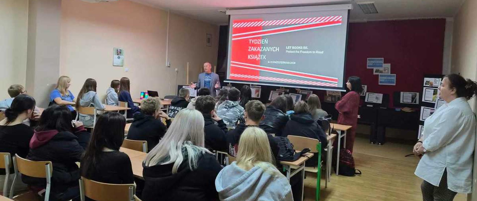 Na zdjęciu widoczna jest prelekcja lub lekcja szkolna zorganizowana w sali lekcyjnej z udziałem młodzieży.
W głębi sali, pośrodku, stoi mężczyzna w średnim wieku, który przemawia do zgromadzonych. Ma na sobie jasną marynarkę, niebieską koszulkę i ciemne spodnie. Za nim, na ścianie, zawieszony jest duży ekran projekcyjny. Wyświetlany jest na nim slajd z czerwono-białymi pasami u góry i u dołu oraz napisami: „TYDZIEŃ ZAKAZANYCH KSIĄŻEK” oraz anglojęzycznym hasłem „LET BOOKS BE. Protect the Freedom to Read”. Poniżej widnieje data wskazująca na październik.
Słuchacze – grupa około dwudziestu nastolatków – siedzą tyłem lub profilem do obiektywu, wypełniając ławki szkolne ustawione w rzędach. Większość z nich ma ciemne ubrania. Na pierwszym planie wyróżniają się dwie dziewczyny o jasnych włosach (jedna z długimi, platynowymi włosami, druga w szarej bluzie z kapturem).
Po prawej stronie sali stoją dwie kobiety. Jedna z nich, odwrócona tyłem, ma na sobie bordową bluzę i ciemne spodnie. Druga, stojąca bliżej prawej krawędzi kadru i zwrócona profilem w lewą stronę, to kobieta w białej koszuli i wzorzystych spodniach, która z uwagą obserwuje spotkanie.
Klasa ma jasne ściany, a na suficie zamontowane są podłużne lampy oraz rzutnik skierowany na ekran. Po prawej stronie, w tle, znajduje się ciemna ściana, na której wiszą tablice z dodatkowymi materiałami i plakatami. Wnętrze jest dobrze oświetlone światłem sztucznym.