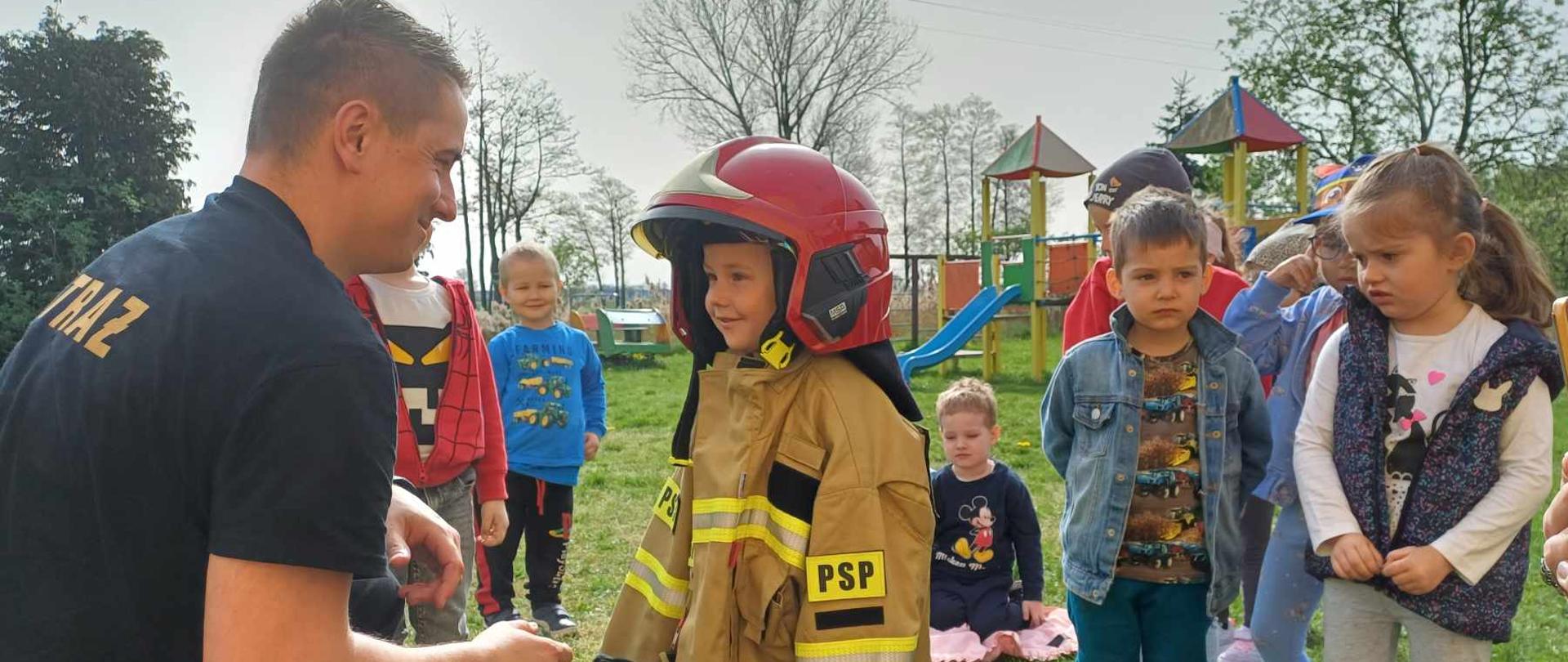 Zdjęcie przedstawia strażaka i dziecko w stroju strażackim, wokół inne dzieci, które obserwują.