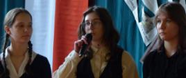 Na zdjęciu występują trzy dziewczyny - uczennice klasy 1A PLSP biorące udział w szkolnej akademii przygotowanej z okazji Święta Narodowego Konstytucji 3 Maja. Uczennice ubrane są odświętnie, w białe koszule i czarne bluzy. Zdjęcie wykonane jest w chwili kiedy jedna z uczennic, ta która jest na środku trzyma w ręce mikrofon i recytuje wiersz. W tle, po lewej stronie zdjęcia widać, w pionie flagę biało-czerwoną. Po prawej stronie w tle, którego kolor jest zielony widać fragment dekoracji wykonanej z gazet. 