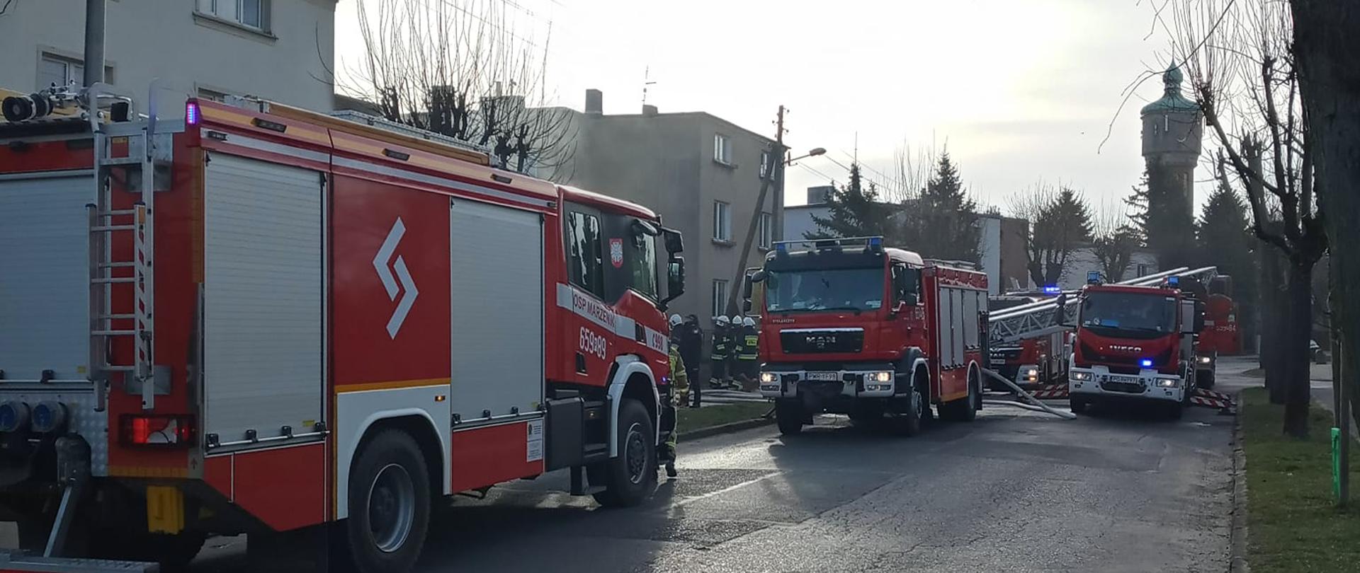 Zdjęcie przedstawia trzy wozy strażackie stojące na jezdni. W tle dwa domy