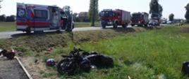 Zdjęcie przedstawia rozbity motocykl znajdujący się w przydrożnym rowie. Na drodze stoi ambulans ZRM oraz samochody pożarnicze z JRG Bochnia: GBARt 2,5/16, SCRt, SLRR.