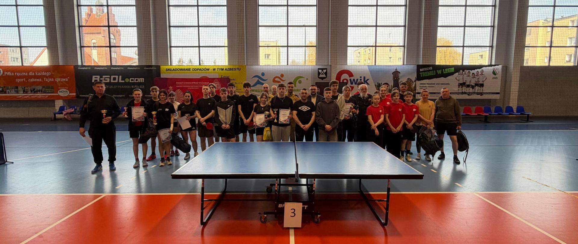 Wspólne zdjęcie grupowe wszystkich uczestników i organizatorów zawodów. Duża grupa osób stoi w rzędzie za stołem do tenisa stołowego z numerem 3, ustawionym na środku hali sportowej. W tle widać duże okna hali, przez które wpada dzienne światło, oraz liczne banery sponsorów i herby.