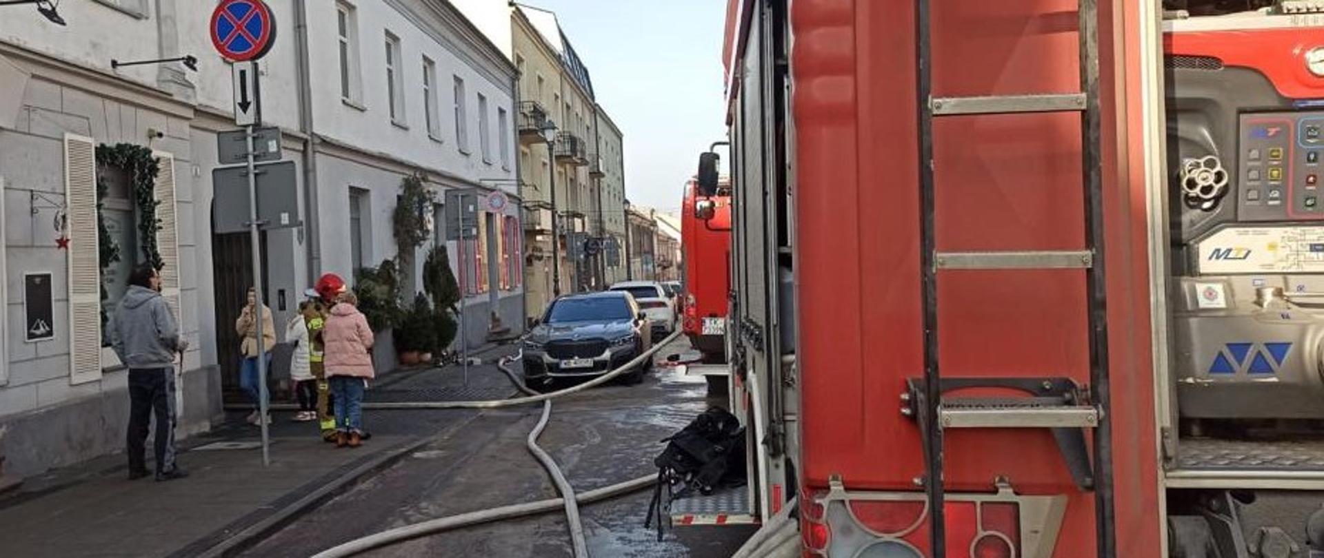 Zdjęcie przedstawia ulicę Świętego Leonarda. Z prawej strony stoi samochód pożarniczy z podpiętymi wężami gaśniczymi, które wchodzą do najbliższej bramy w pierzei kamienic znajdujących się po lewej stronie. Przy bramie widać ratownika wśród kilku obserwatorów akcji ratowniczej.