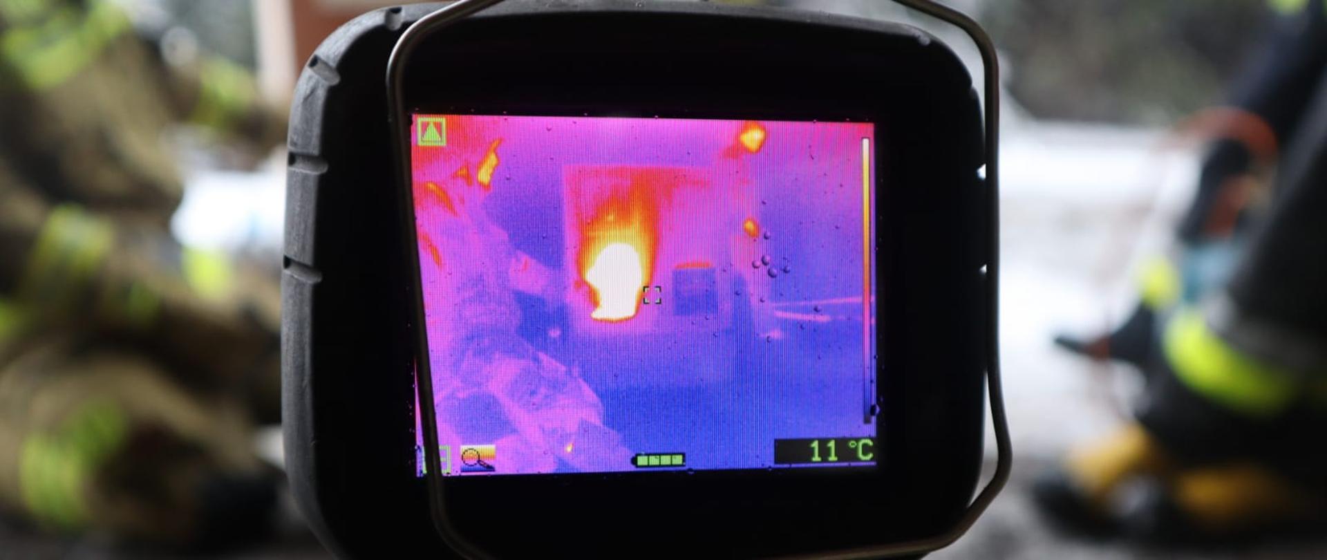 Zdjęcie przedstawi widok z kamery termowizyjnej palącego się obiektu w tle zamazane osoby strażaków