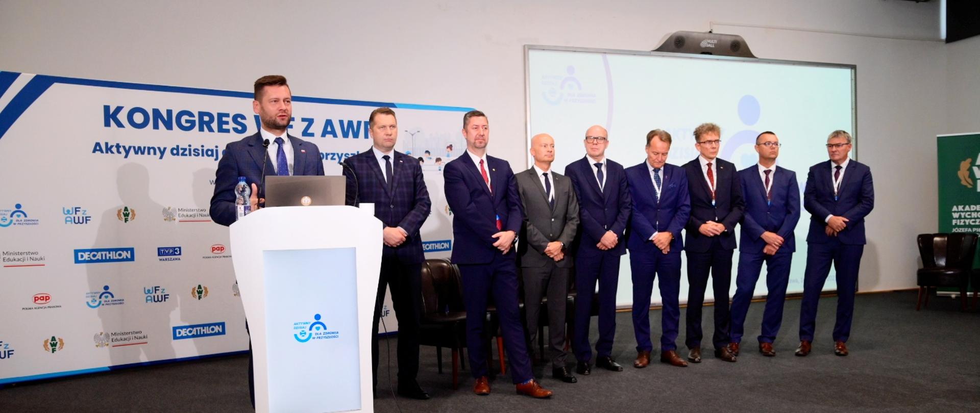 Na sali pod ścianką z napisem Kongres WF z AWF stoi grupa mężczyzn w garniturach, przed nimi minister Bortniczuk stoi za mównicą i mówi do mikrofonu.