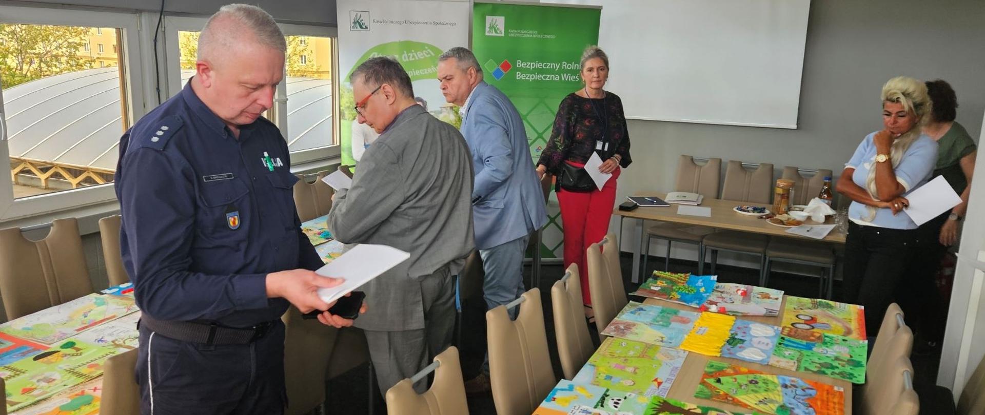 W pokoju konferencyjnym trzy kobiety i trzech mężczyzn, jeden z nich mundurze policyjnym, stoi z notatkami w rękach przy dwóch stołach, na których leży kilkadziesiąt prac plastycznych dzieci. 