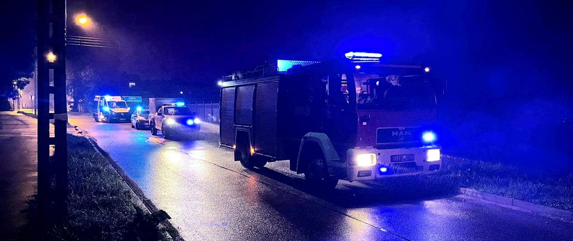 Obraz przedstawia pojazdy pożarnicze z włączonymi sygnałami alarmowymi świetlnymi stojące wzdłuż jezdni