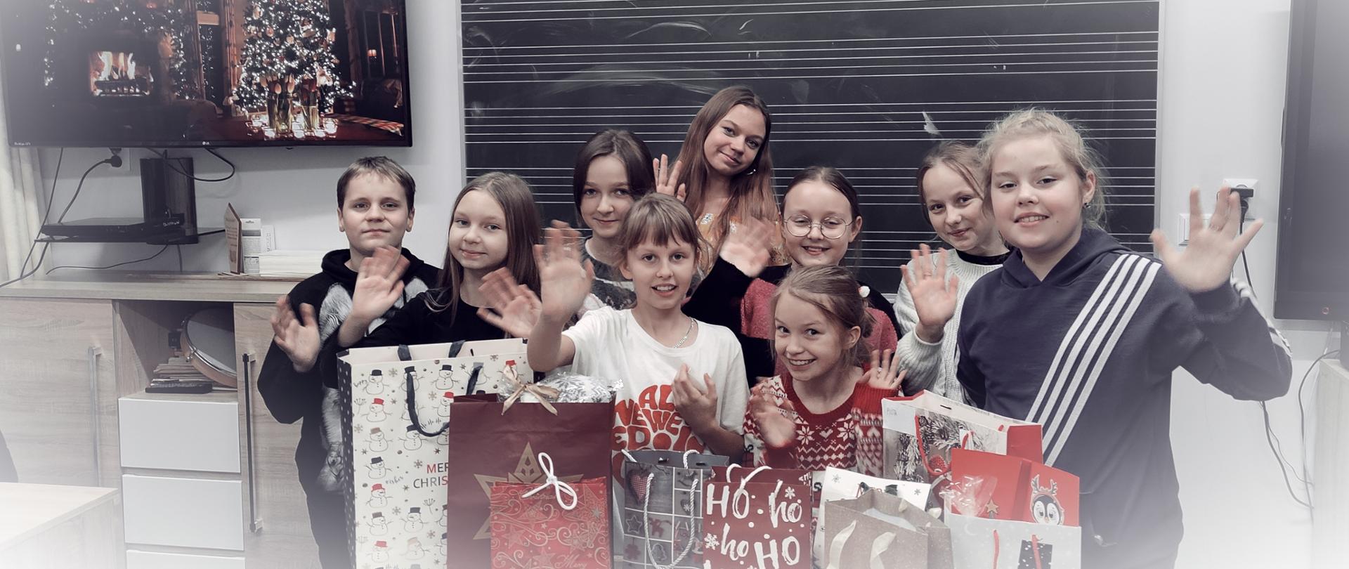 Grupa uczniów stoi na tle tablicy w pięciolinie. Na ławce przed uczniami znajdują się świąteczne torby z prezentami mikołajkowymi. Uczniowie uśmiechają się i machają rękami na powitanie.