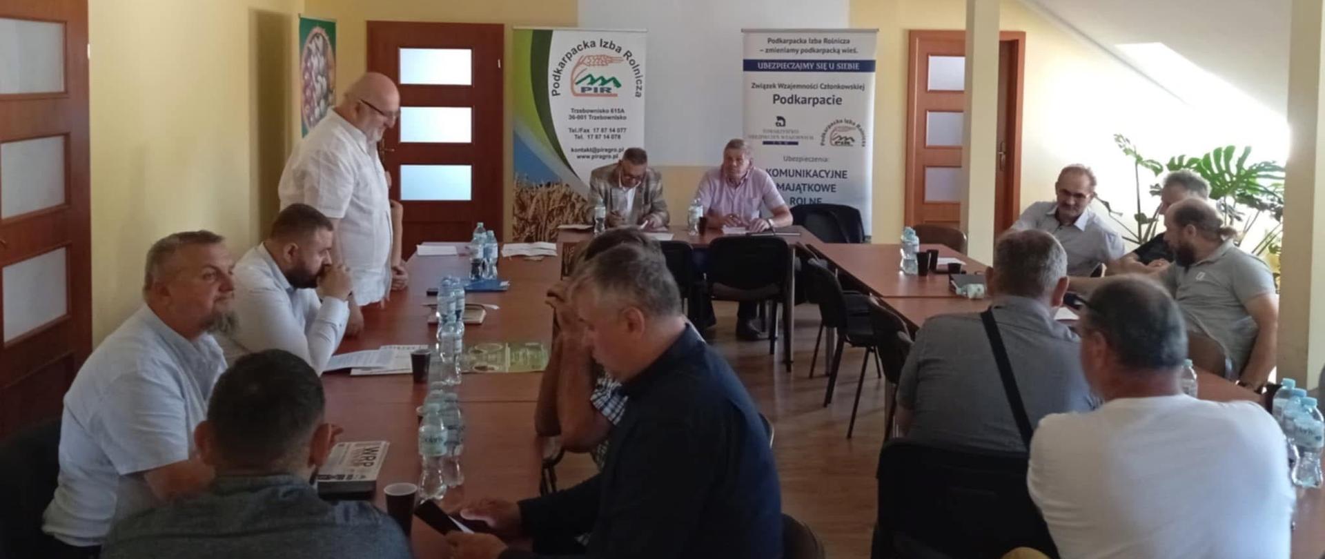 Grupa mężczyzn w jasnym pomieszczeniu. Siedzą przy stołach ustawionych w kształcie litery ,,U''. Jeden z mężczyzn stoi i mówi.