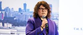 Dyrektor Generalna Ewa Weber podczas VI Krajowego Kongresu Forów Skarbników, trzyma w ręku mikrofon, za nią ścianka ze zdjęciem panoramy Warszawy.