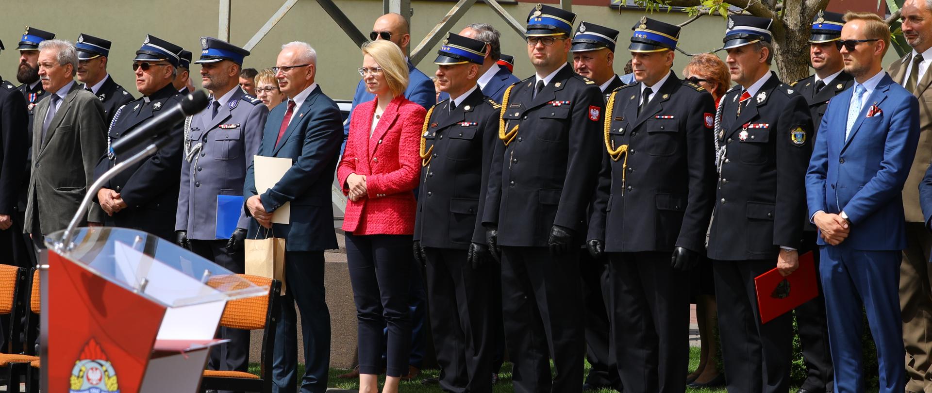 grupa ludzi stoi w szeregu, wśród nich strażacy, przed nimi mównica, w tle budynek żółto brązowy
