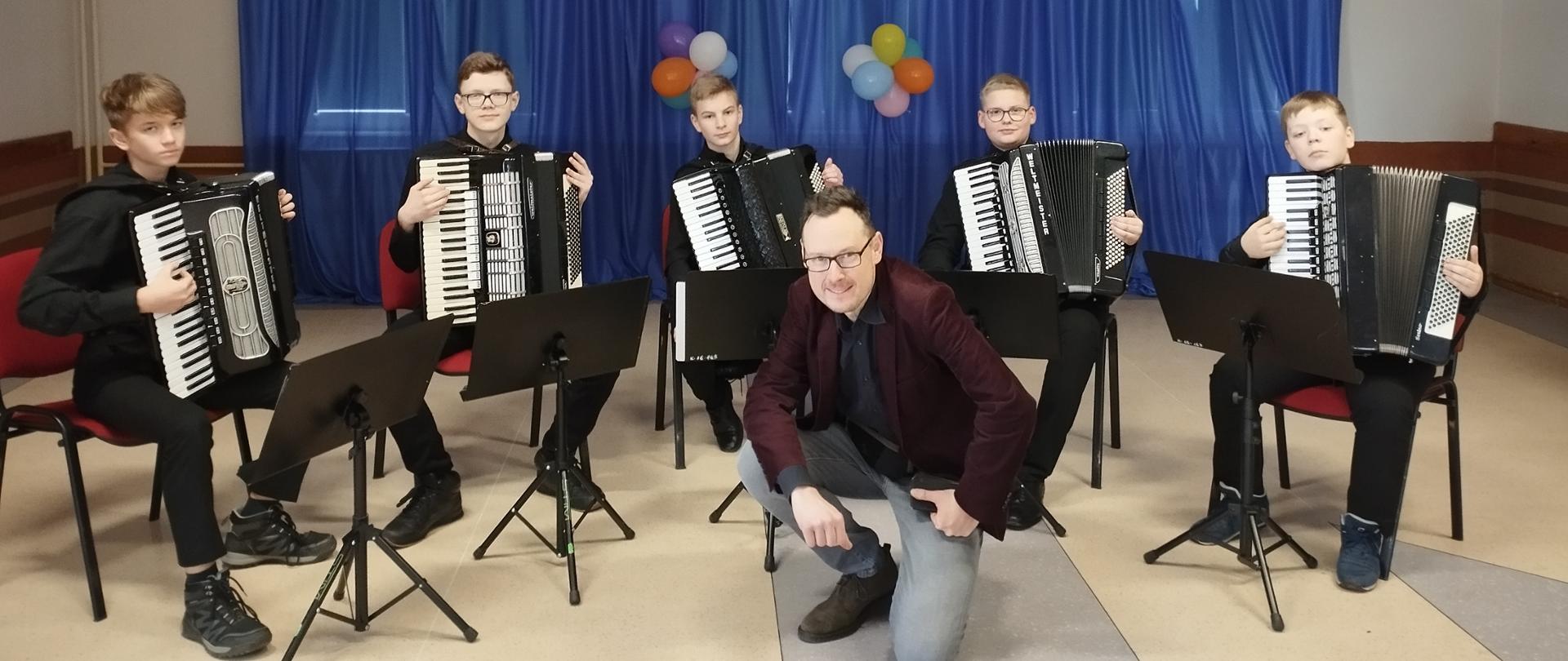 Uczniowie kwintetu akordeonowego siedzą w półkolu przy pulpitach pozując do zdjęcia. Przed nimi w pozycji półklęczącej pozuje nauczyciel Ryszard Borysionek. W tle zasłona w kolorze granatowym z przypiętymi literami tworzącymi tekst: 10-te urodziny wejherowskiego stowarzyszenia na rzecz osób z autyzmem.