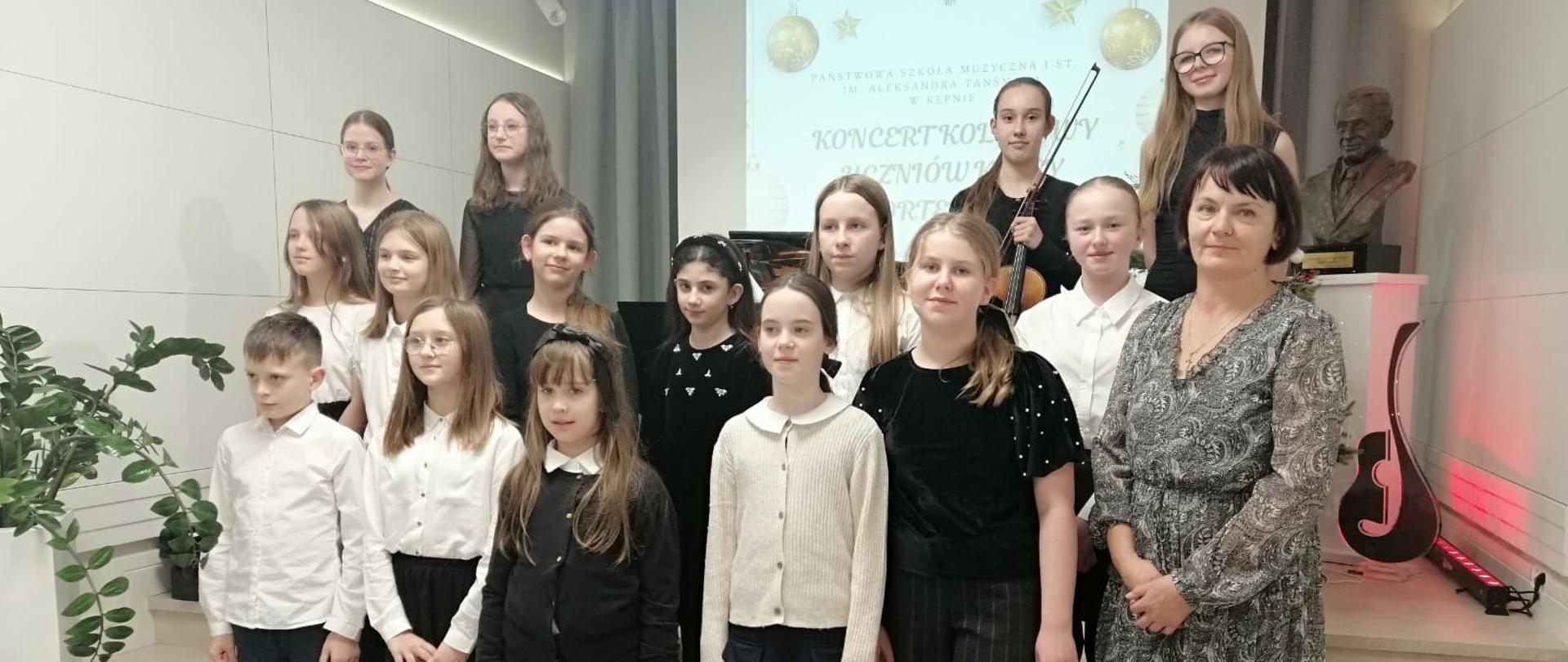 Zdjęcie przedstawia grupę uczniów z nauczycielką. Wszyscy ubrani są elegancko w odcieniach bieli, czerni i granatu. Kilka uczennic trzyma instrumenty, m.in. skrzypce i flet poprzeczny. Grupa stoi na schodkach sceny we wnętrzu nowoczesnej sali koncertowej. W tle znajduje się projektor z napisem: „Koncert kolędowy uczniów fortepianu”. Dekorację dopełnia świąteczna grafika z bombkami, fortepian, popiersie kompozytora oraz ozdobna gitara podświetlona czerwonym światłem. Obok sceny widać także dużą doniczkową roślinę. 