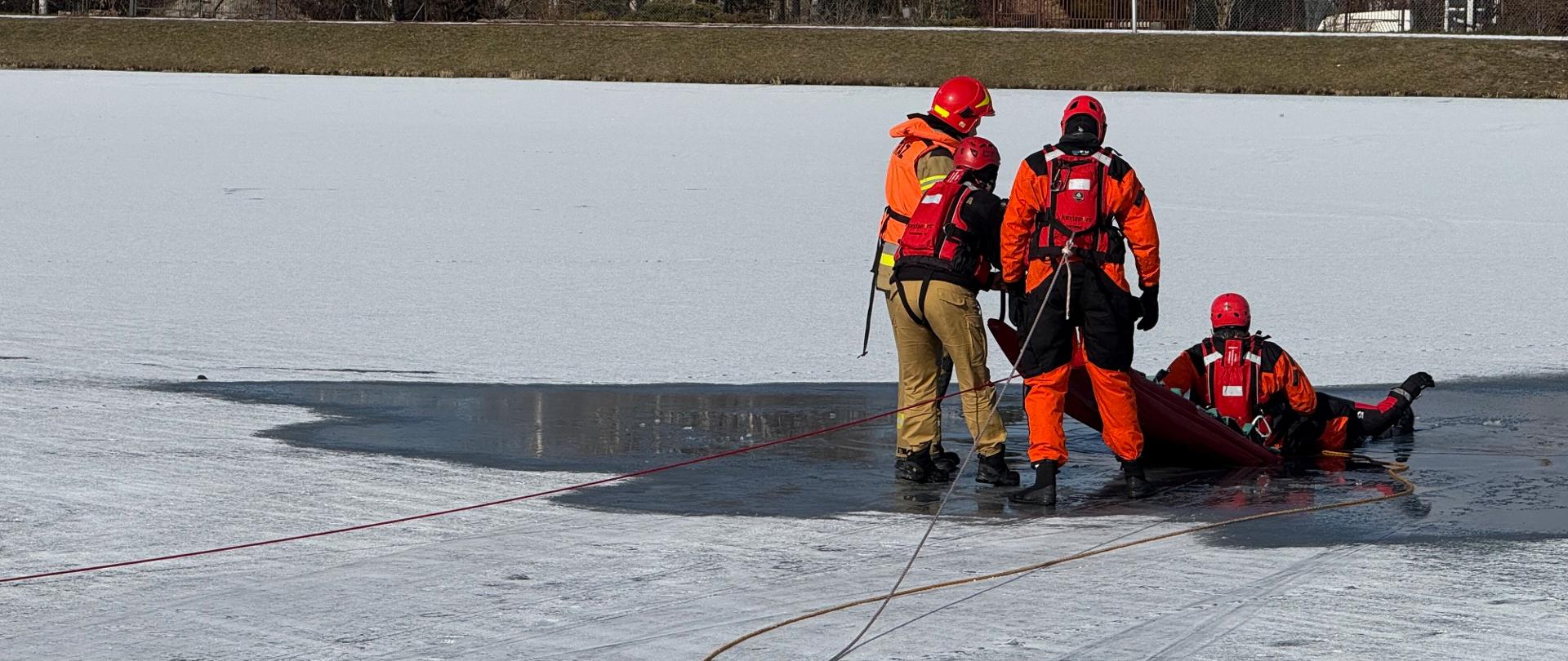 Zdjęcie przedstawia trzech strażaków prowadzących ćwiczenia na zamarzniętym zbiorniku wodnym. Dwóch strażaków, ubranych w pomarańczowe kombinezony ratunkowe i kaski, stoi na lodzie. Jeden z nich, bliżej centrum kadru, jest pochylony nad trzecim strażakiem, który leży na saniach ratowniczych, częściowo zanurzony w wodzie. Wokół nich rozłożone są liny.
W tle, po drugiej stronie zamarzniętego jeziora, widoczne są domy i drzewa. Niebo jest błękitne z białymi chmurami. Lód jest popękany w miejscu, gdzie odbywa się ćwiczenie.