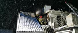 Na zdjęciu budynek mieszkalny z białą elewacją i widocznym fragmentem dachu krytego niebieską blachodachówką. Noc, widoczne opady śniegu z deszczem. Strażacy wchodzący do wnętrza poddasza przez oderwany fragment poszycia dachu. Z prawej strony widoczny fragment drabiny strażackiej oparty o balkon budynku.