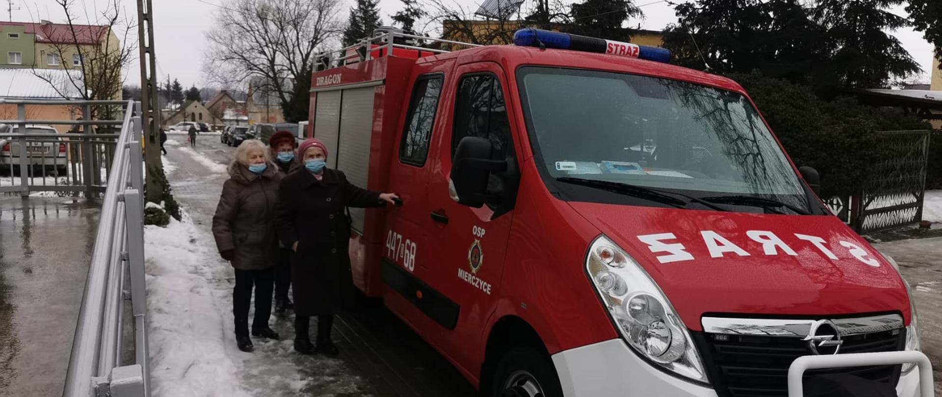 Obraz przedstawia samochód pożarniczy. Przy samochodzie znajduje się troje seniorów których strażacy transportują na szczepienia. W tle droga gminna i drzewa.