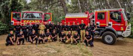 Strażacy pozują do zdjęcia grupowego w środku lasu, za nimi znajdują się wozy strażackie francuskiej straży pożarnej.