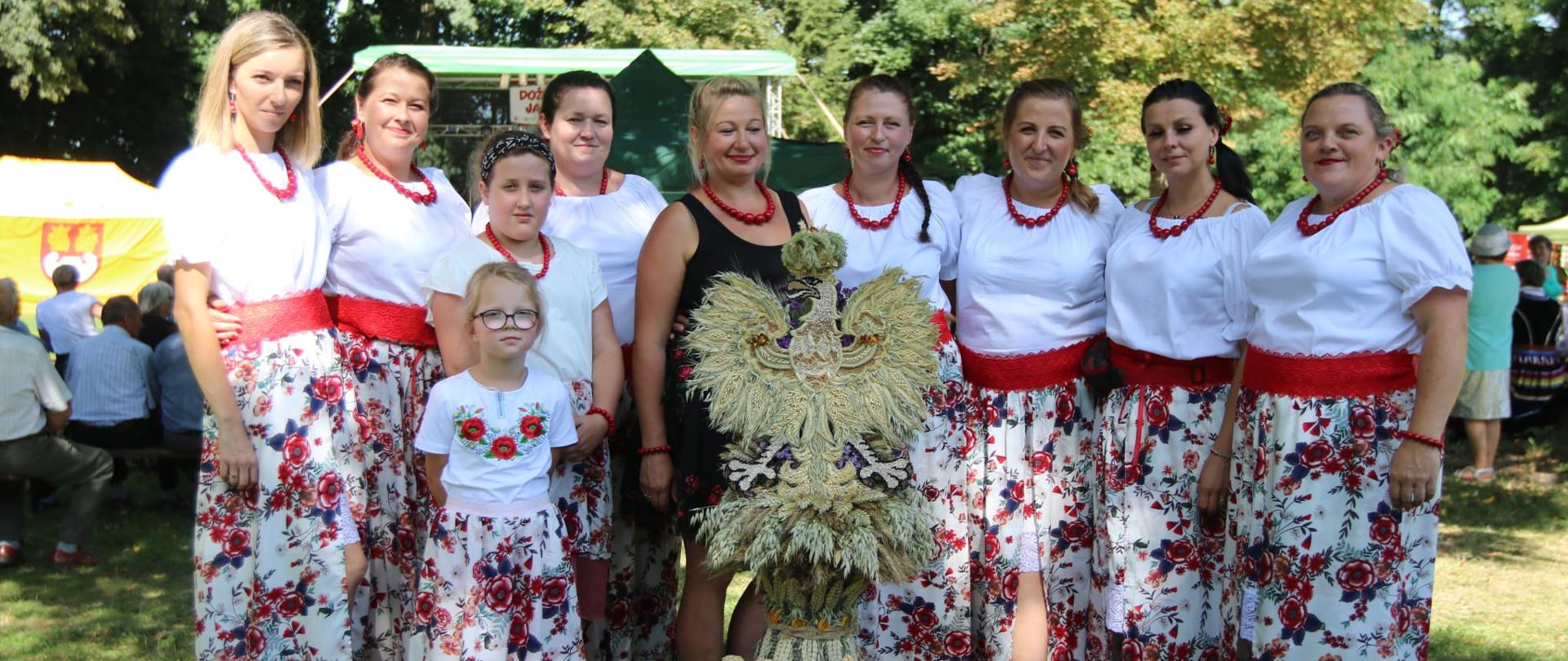 Kobiety i dziewczęta ubrane w stroje regionalne przed nimi wieniec dożynkowy