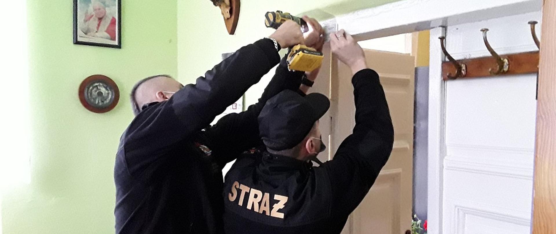 Zdjęcie przedstawia dwóch strażaków z JRG Koluszki montujących czujkę czadu na futrynie drzwi w budynku mieszkalny jednorodzinnym