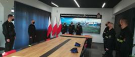Zdjęcie przedstawia strażaków stojących w sali konferencyjnej na uroczystości wręczenia awansów na wyższe stopnie służbowe. Awansowani stoją na wprost w mundurach galowych wraz z komendantem. 