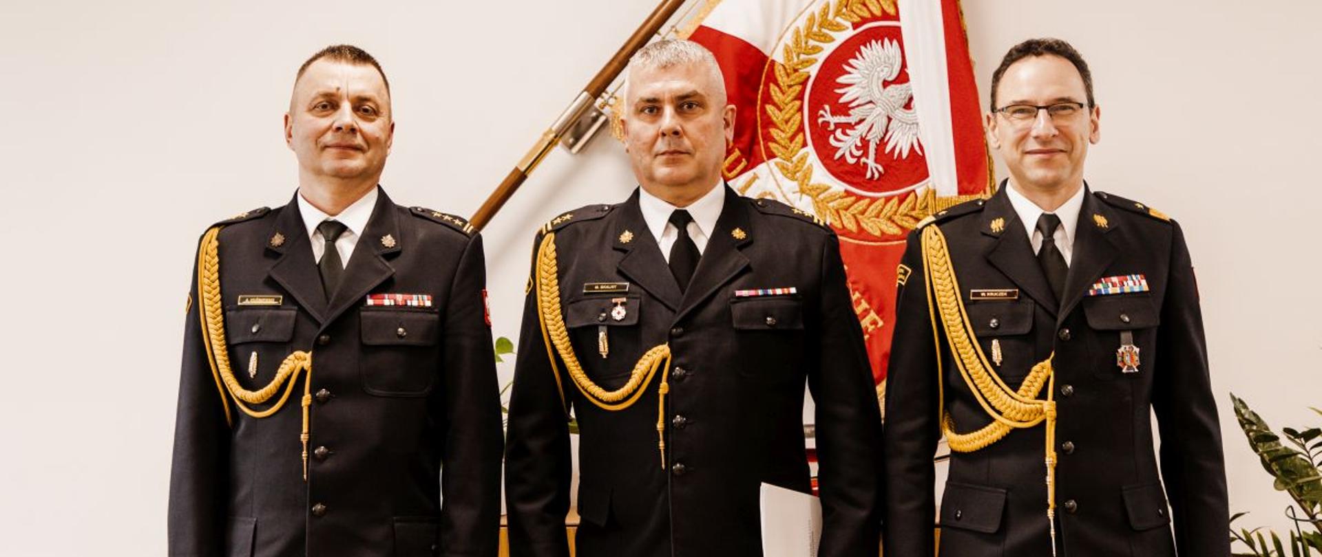 Trzech oficerów Państwowej Straży Pożarnej w galowych mundurach stoi obok siebie na tle czerwono‑białego sztandaru z orłem. Centralnie ustawiony funkcjonariusz trzyma w dłoniach dokument powołania. Obok niego stoją wysocy rangą przedstawiciele PSP, uczestniczący w uroczystości. Zdjęcie dokumentuje oficjalne ogłoszenie zmian kadrowych w województwie opolskim, podczas których bryg. Marcin Skalny otrzymał decyzję o powołaniu na stanowisko Zastępcy Opolskiego Komendanta Wojewódzkiego PSP.