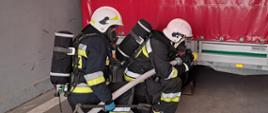 Strażacy OSP w pełnym umundurowaniu specjalnym i sprzęcie izolującym drogi oddechowe. W pozycji przyklęku na kolano z linią gaśniczą i prądownicą.