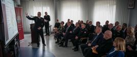 Zdjęcie zrobione w pomieszczeniu strażnicy. W centralnym miejscu pomieszczenia Komendant Powiatowy Państwowej Straży Pożarnej w Lublińcu st. bryg. Janusz Bula przedstawia sprawozdanie z działalności Komendy Powiatowej PSP w Lublińcu za rok 2022 przy pomocy wyświetlanej prezentacji multimedialnej. Naprzeciw Komendanta Powiatowego PSP w Lublińcu zaproszeni goście w pozycji siedzącej wsłuchują się w słowa przedstawianego przedmiotowego sprawozdania. Za prelegentem można zaobserwować baner Komendy Powiatowej PSP w Lublińcu oraz Zastępcę Komendanta Powiatowego PSP w Lublińcu mł. bryg Daniela Kozioł.
