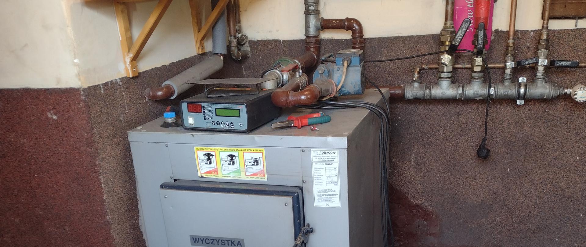 Na zdjęciu widać piec centralnego ogrzewania. Piec w pomieszczeniu piwnicznym. Na piecu sterownik pieca. Wokół pieca instalacja połączeniowa.