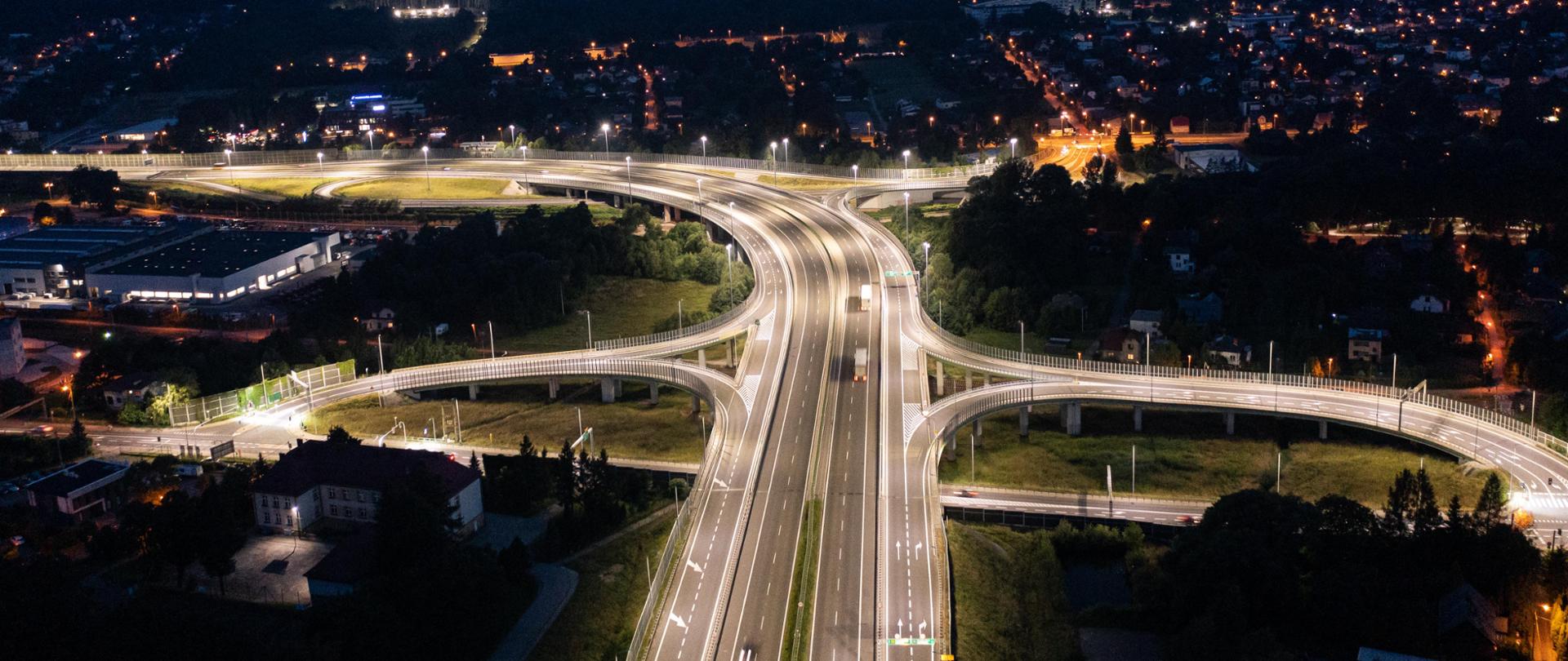 Widok z lotu ptaka na miasto w trakcie zmierzchu, ukazujący złożoną sieć oświetlonych dróg i autostrad. Światła tworzą żywy kontrast z ciemniejącym niebem, podkreślając ruch uliczny i złożoność krajobrazu miejskiego. Ten obraz jest interesujący, ponieważ pokazuje infrastrukturę i organizację systemu transportu miejskiego oraz perspektywę aktywności ludzkiej w okresie przejścia między dniem a nocą.