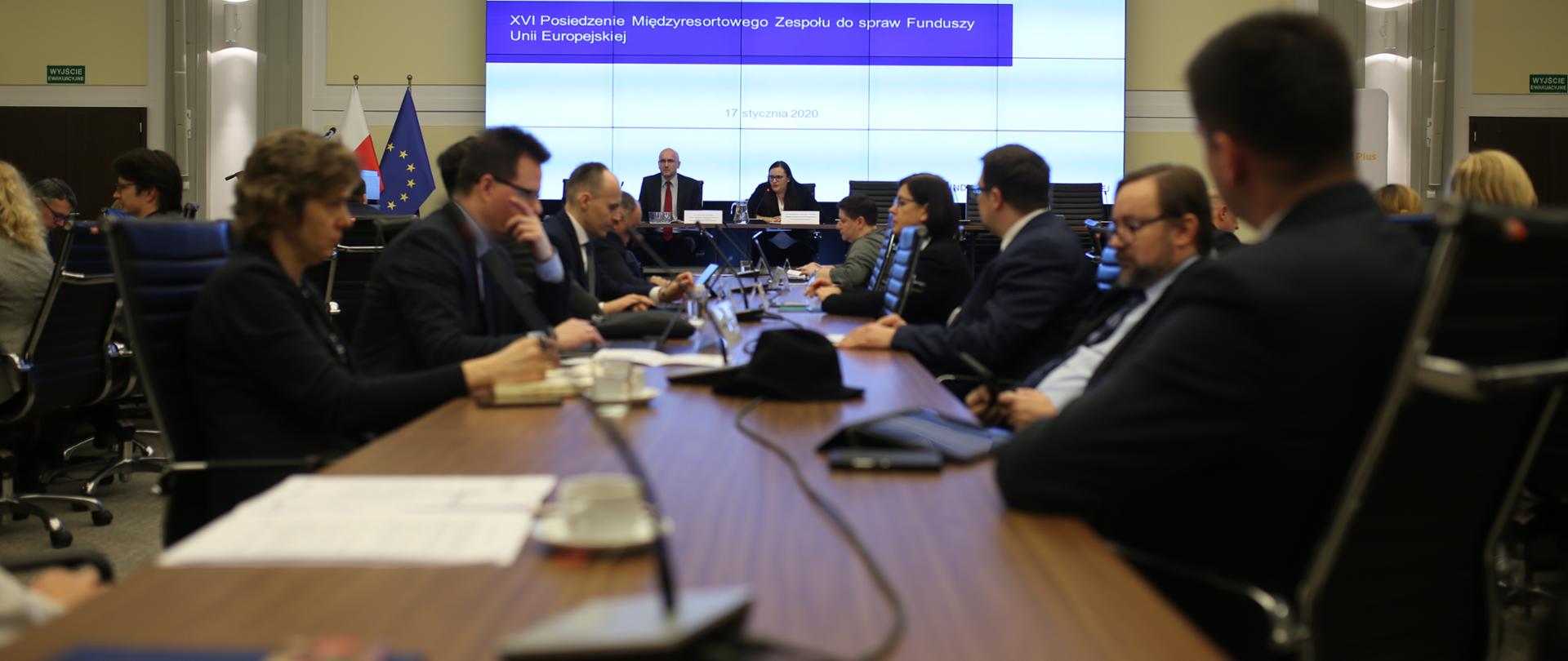 Na zdjęciu sala konferencyjna, na pierwszym planie stół, przy nim siedzą uczestnicy spotkania, w tle stół prezydialny, przy nim siedzi minister Małgorzata Jarosińska-Jedynak.