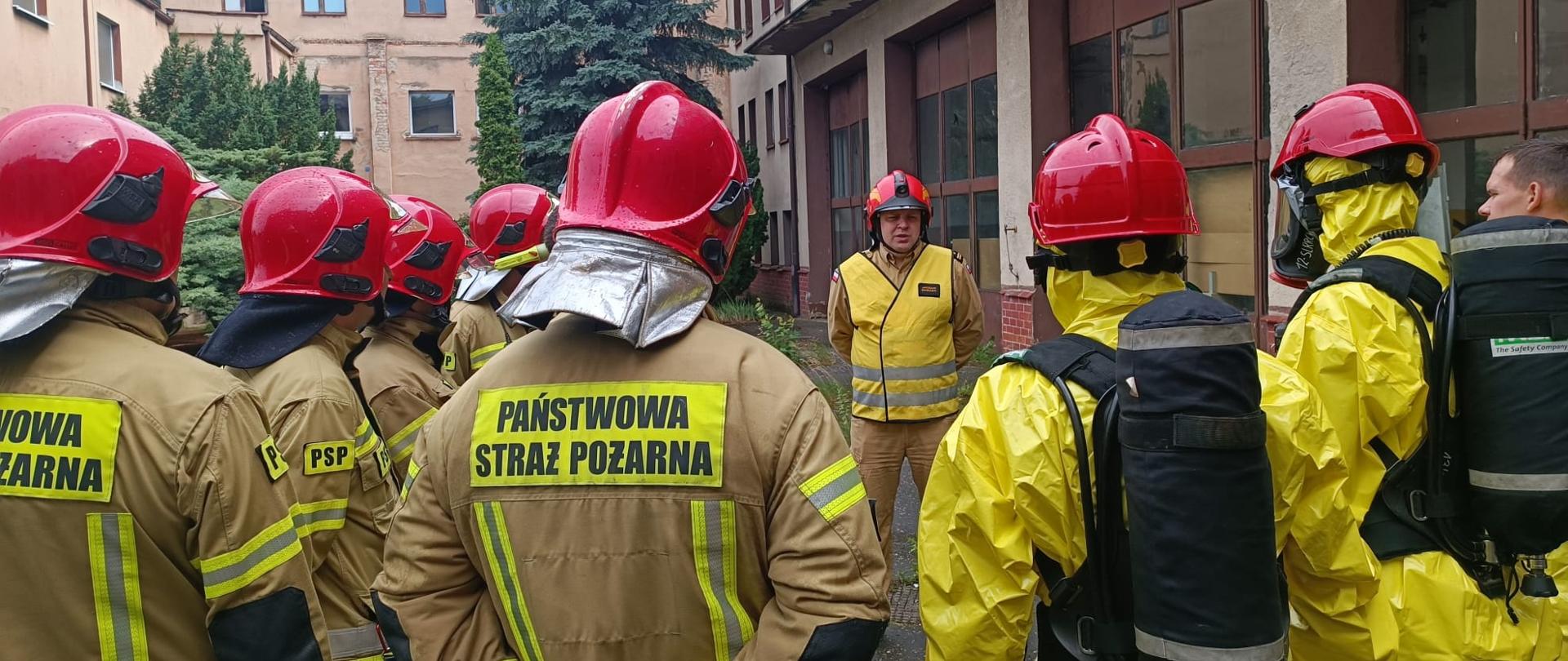 grupa strażaków w jasnych mundurach bojowych i w czerwonych hełmach stoi w okręgu, jeden strażak w żółtej kamizelce i czerwonym hełmie mówi di nich, w tle duży budynek