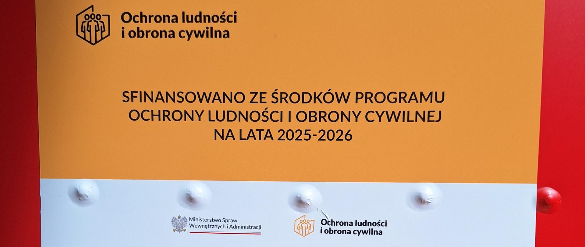 dwóch części:
• Po lewej stronie znajduje się pomarańczowy panel z czarnym tekstem „Ochrona ludności i obrona cywilna” oraz logo przypominającym znak systemu ochrony ludności.
• Po prawej stronie widoczna jest biała część z informacją, że projekt został sfinansowany ze środków Programu Ochrony Ludności i Obrony Cywilnej na lata 2025–2026. Pod spodem znajduje się nazwa Ministerstwo Spraw Wewnętrznych i Administracji oraz adres strony internetowej.
Całość wygląda jak oficjalna tablica informująca o dofinansowaniu lub modernizacji elementu infrastruktury związanej z ochroną ludności.
