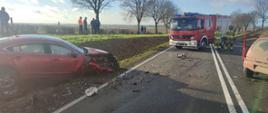 Po lewej stronie zdjęcia widać rozbity samochód osobowy w rowie. Po środku na drodze stoi samochód pożarniczy, przed nim dwóch strażaków. Po prawej widać tył drugiego rozbitego samochodu