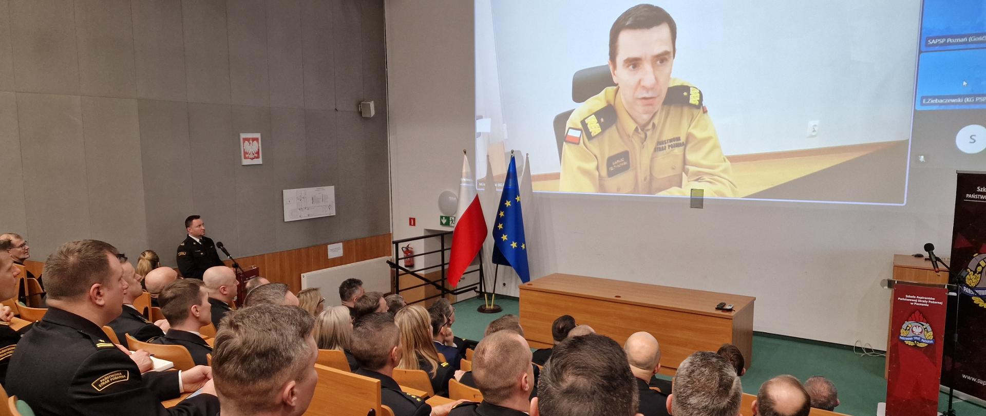 Na auli siedzą funkcjonariusze PSP i osoby cywilne podczas narady, na ekranie wyświetlony wizerunek komendanta głównego PSP biorącego udział on-line w naradzie