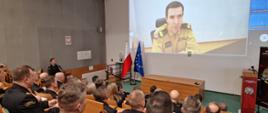 Na auli siedzą funkcjonariusze PSP i osoby cywilne podczas narady, na ekranie wyświetlony wizerunek komendanta głównego PSP biorącego udział on-line w naradzie