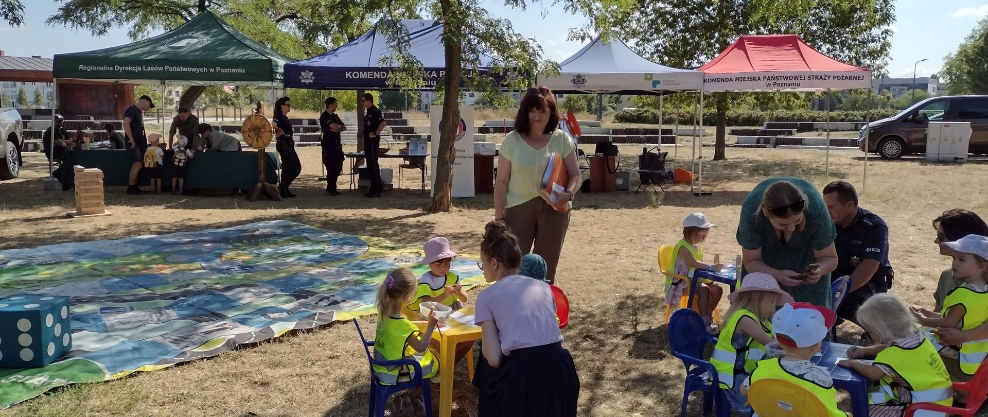 : Dzieci siedzą przy stoliczkach rozstawionych na trawie i kolorują obrazki. W te namioty organizatorów festynu.