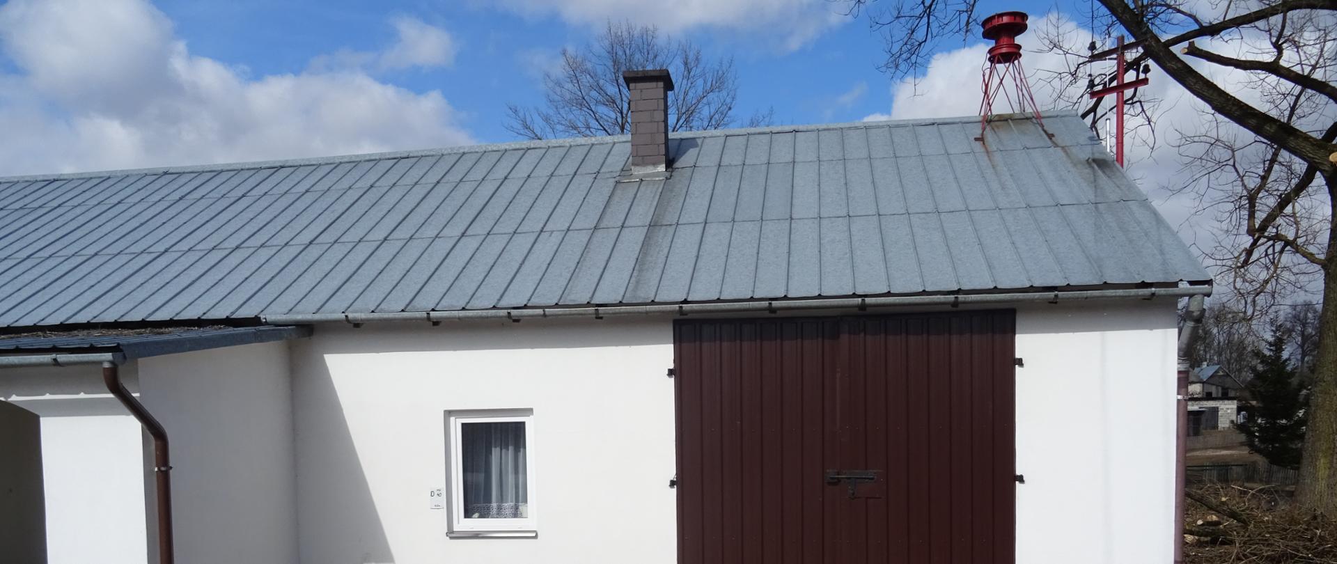 Zdjęcie przedstawia strażnicę OSP Stoki budynek parterowy o białych ścianach i spadzistym dachu na dachu znajduje się syrena alarmowa