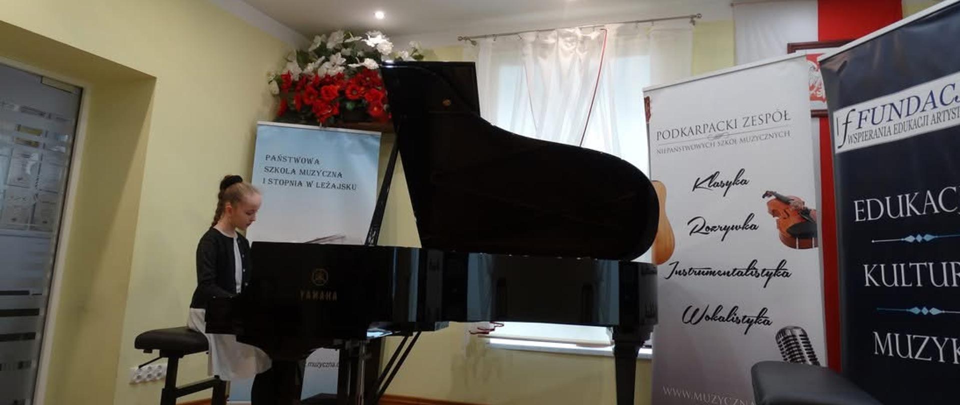 Uczennica w czasie prezentacji konkursowej gra na fortepianie. Obok widoczne oraz rollupy organizatora oraz dekoracja kwiatowa